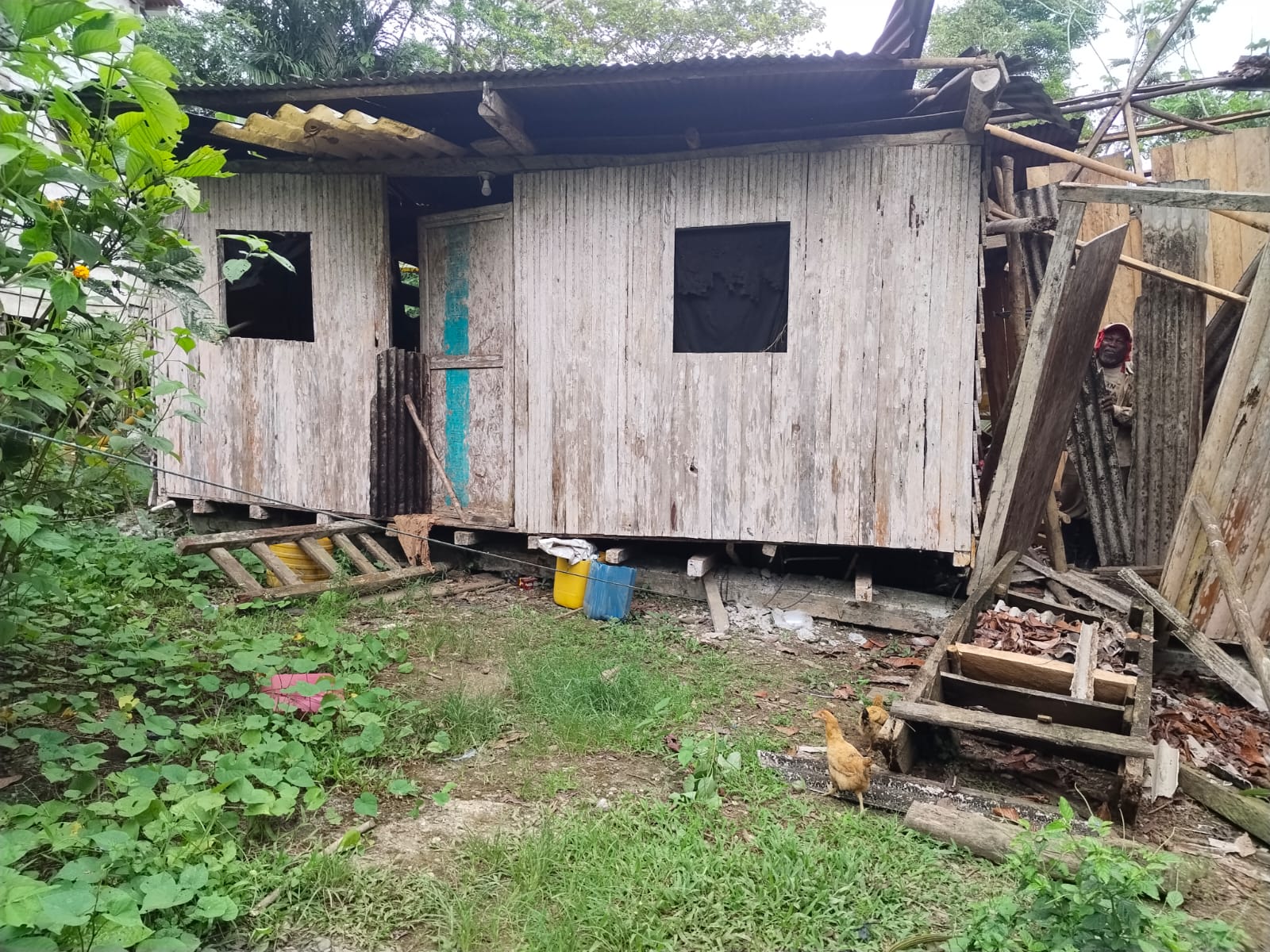 Casa de tablas descuadrada sobre sus bases, con el techo de zinc suelto y una persona al lado